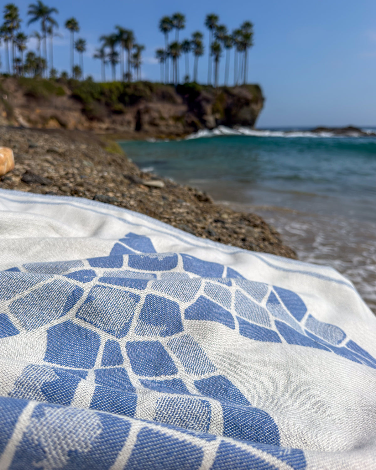 A close-up of an organic Turkish cotton beach towel with a blue geometric mosaic pattern, laid out on a beach with ocean water, rocky shore, and palm trees in the background.