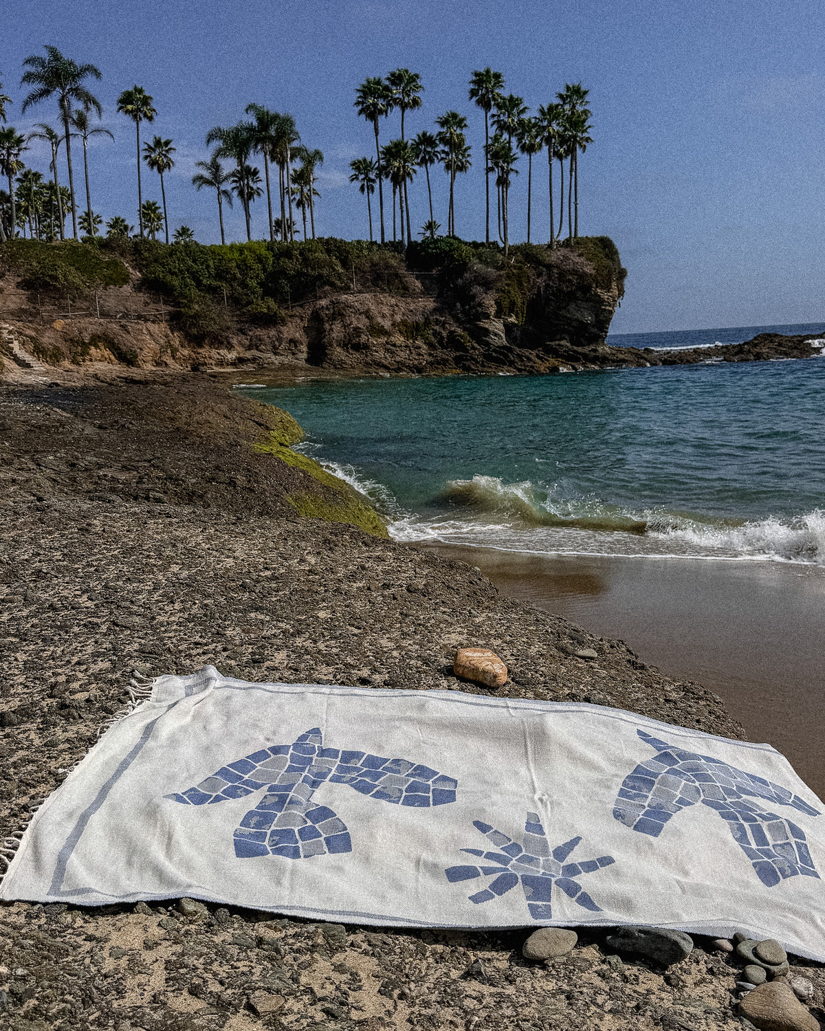 An organic Turkish cotton beach towel with blue mosaic-style bird and sun motifs lies on a rocky beach in front of a tropical coastline with palm trees, blue water, and a clear sky.