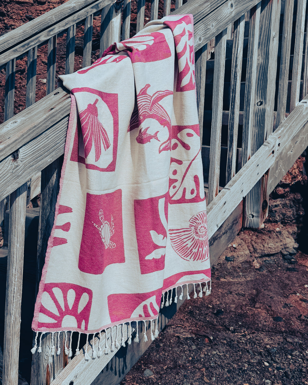 An organic beach towel with a cream base and pink geometric and animal motifs, draped over a weathered wooden staircase railing outdoors.