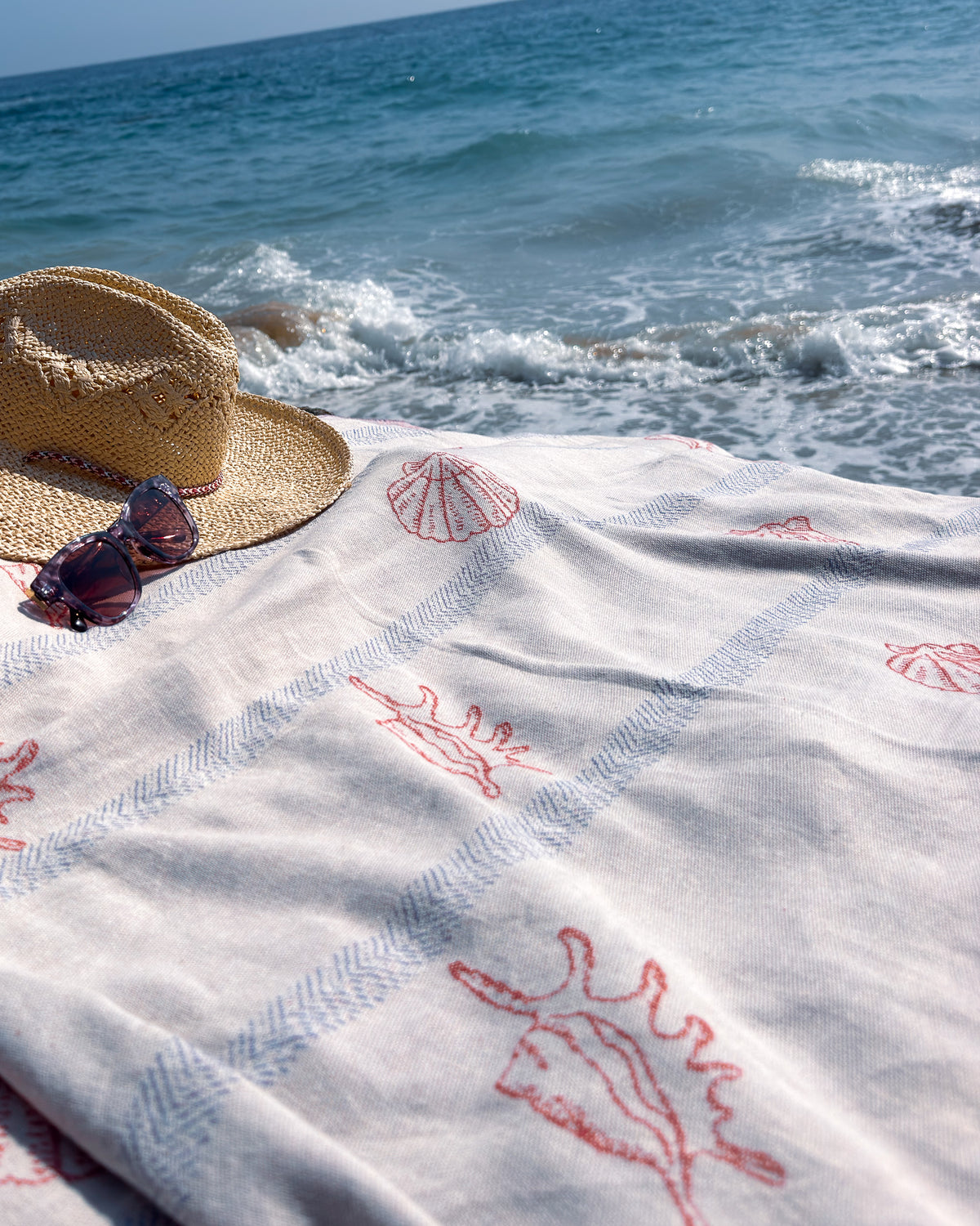 An organic turkish cotton beach towel with red seashell and blue stripe patterns is spread out on a sunny beach, styled with a straw hat and sunglasses in front of the blue ocean.