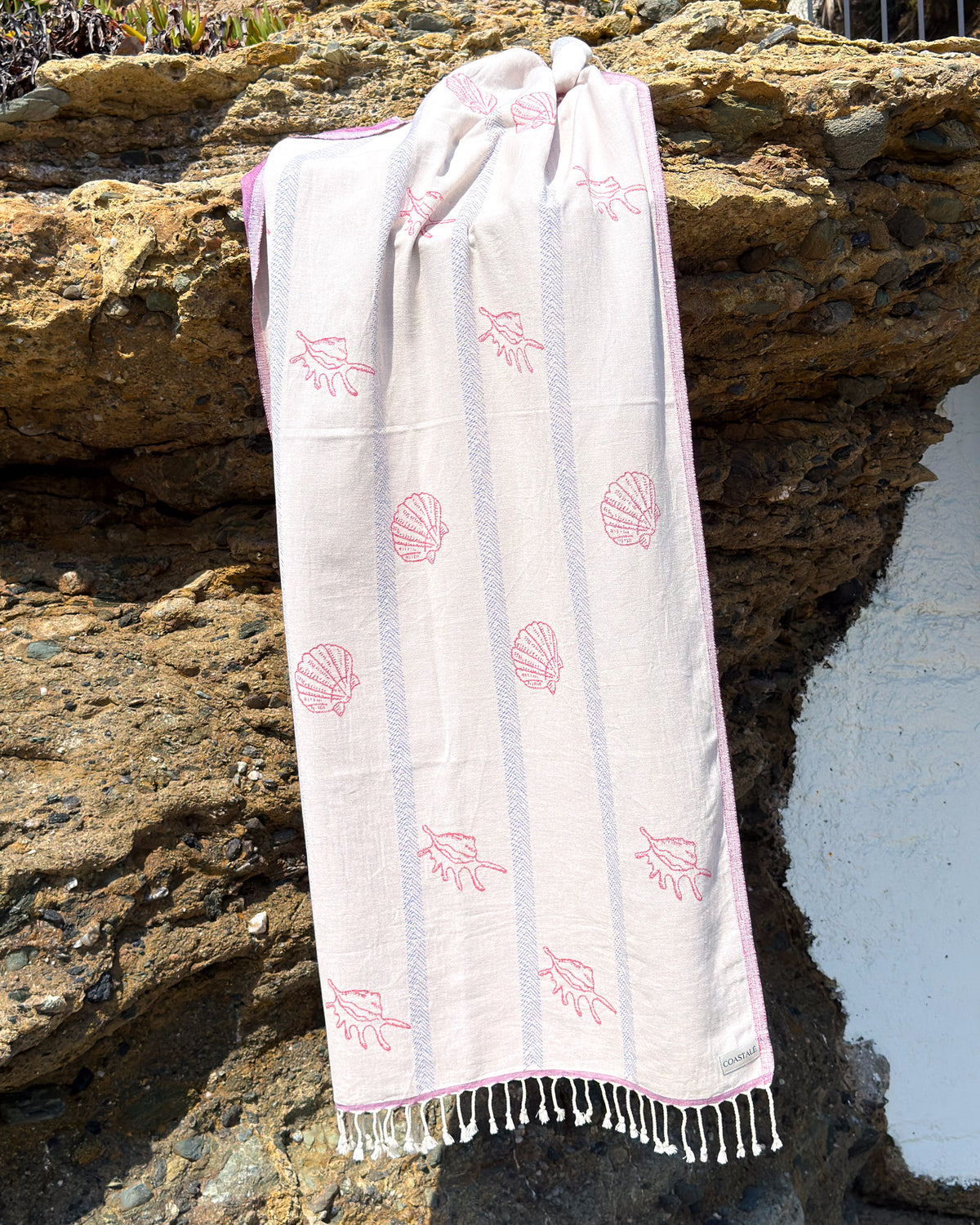 An organic turkish cotton beach towel with seashell and starfish patterns, draped over a rocky outdoor wall with white stucco visible on the right.