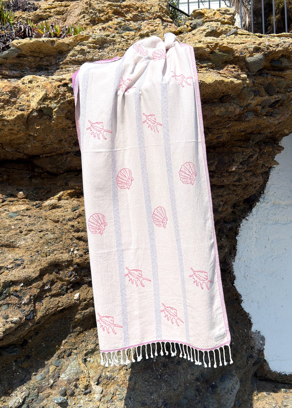 An organic turkish cotton beach towel with seashell and starfish patterns, draped over a rocky outdoor wall with white stucco visible on the right.