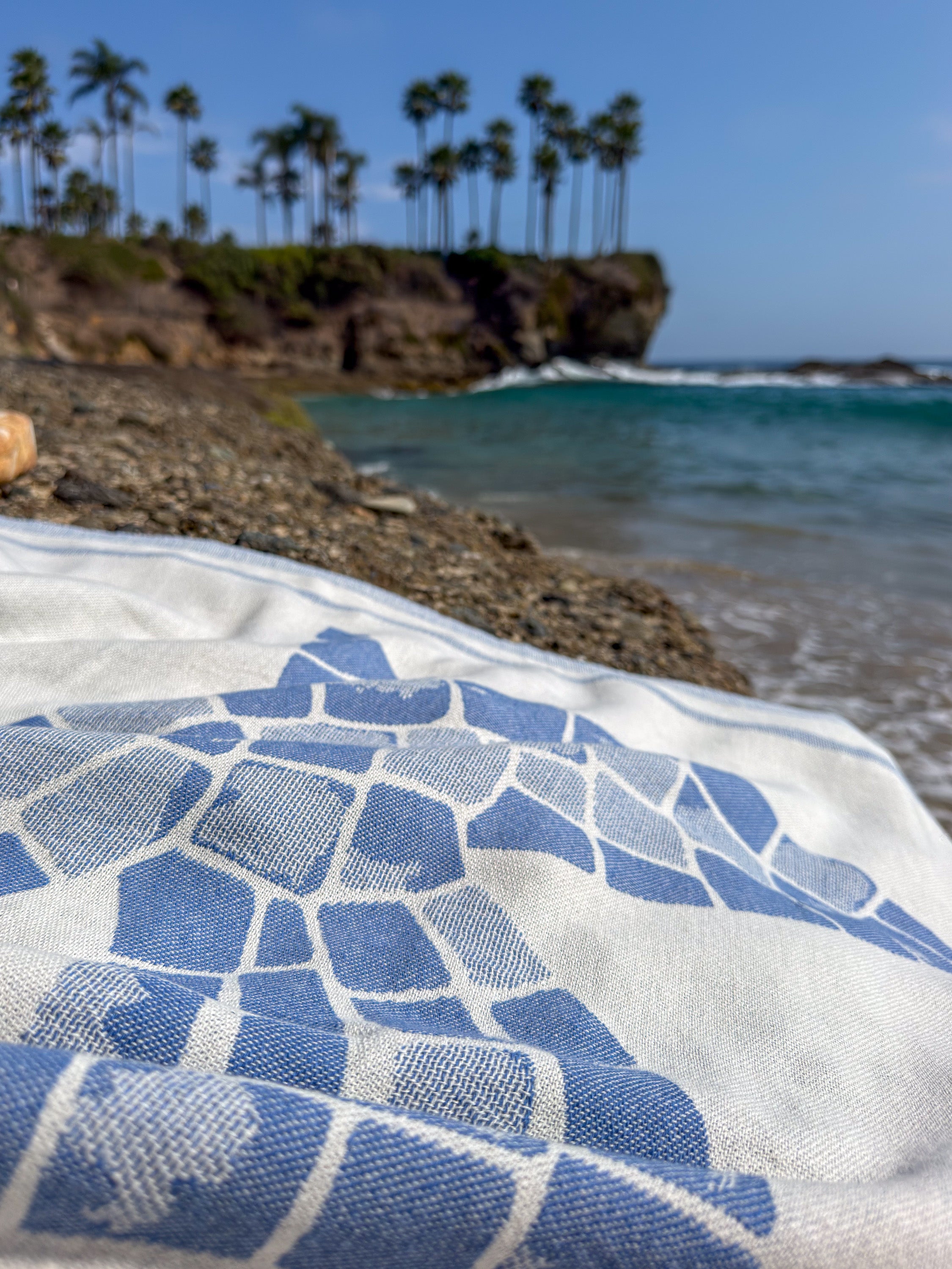 A close-up of an organic Turkish cotton beach towel with a blue geometric mosaic pattern, laid out on a beach with ocean water, rocky shore, and palm trees in the background.