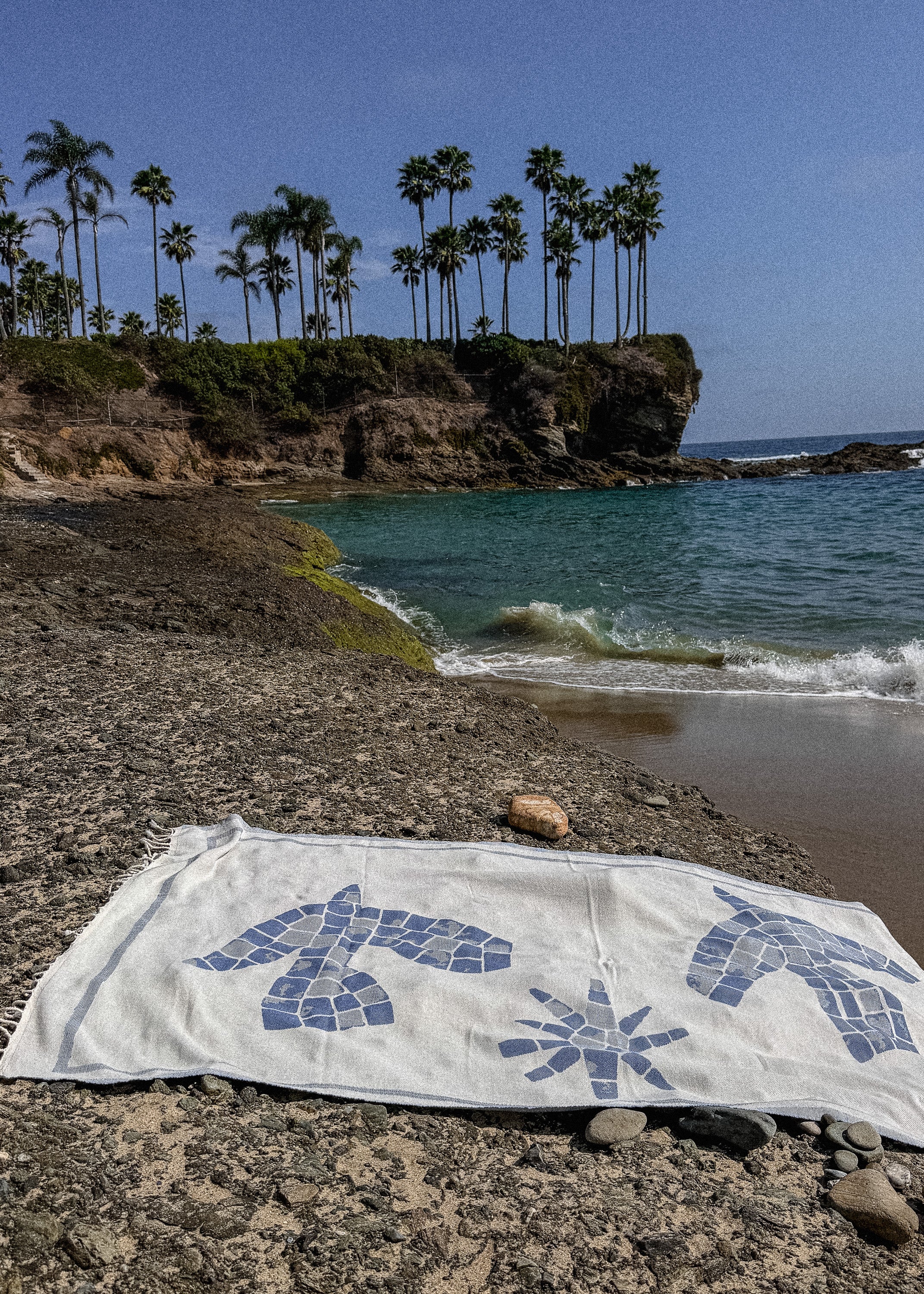 An organic Turkish cotton beach towel with blue mosaic-style bird and sun motifs lies on a rocky beach in front of a tropical coastline with palm trees, blue water, and a clear sky.