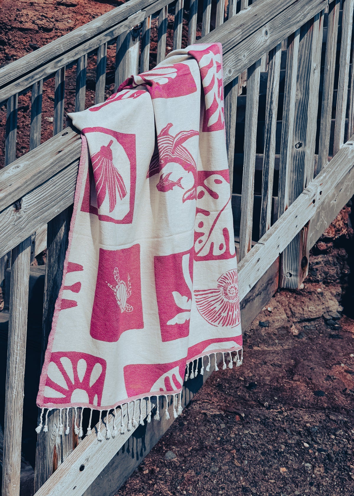 An organic beach towel with a cream base and pink geometric and animal motifs, draped over a weathered wooden staircase railing outdoors.