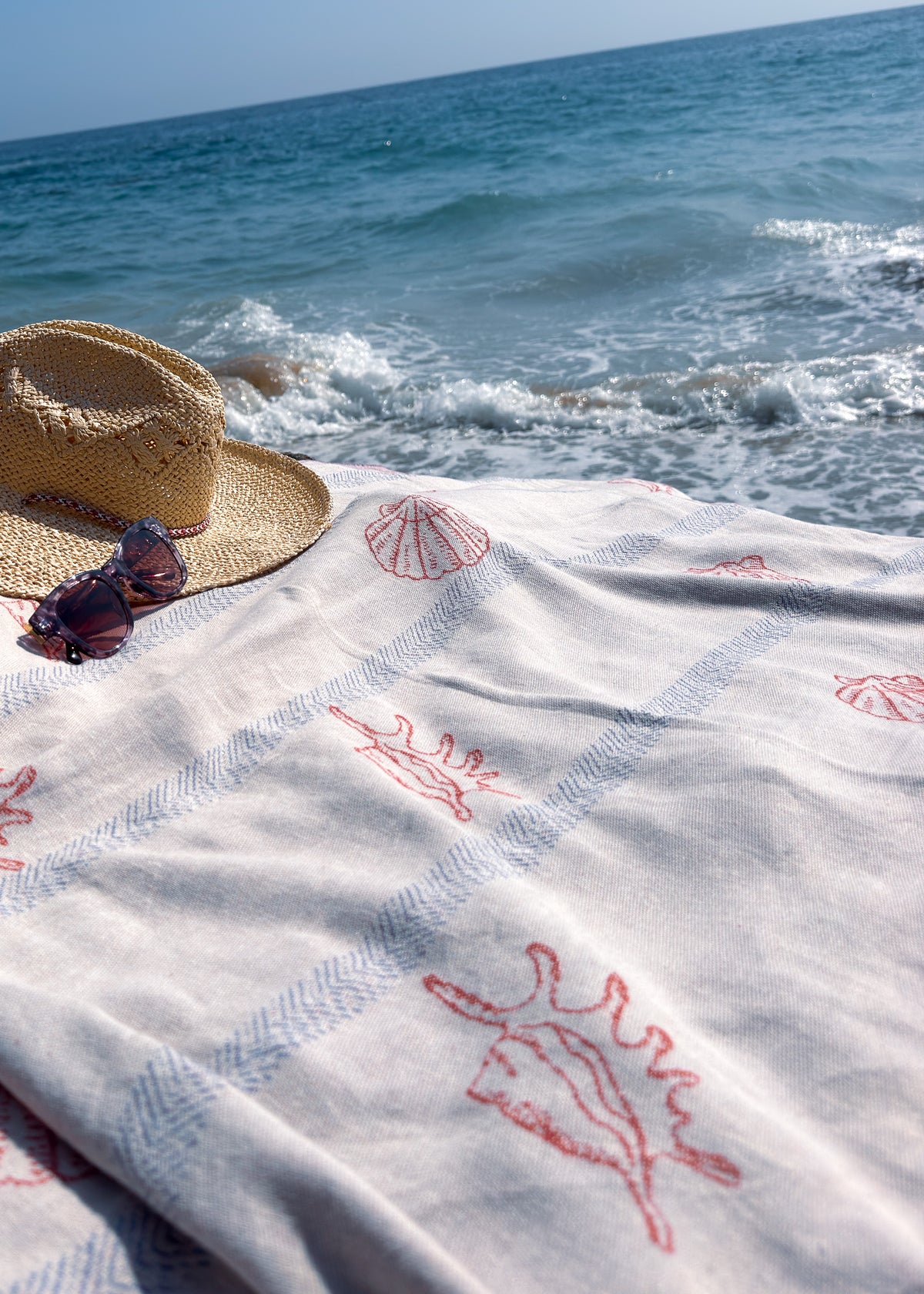 An organic turkish cotton beach towel with red seashell and blue stripe patterns is spread out on a sunny beach, styled with a straw hat and sunglasses in front of the blue ocean.